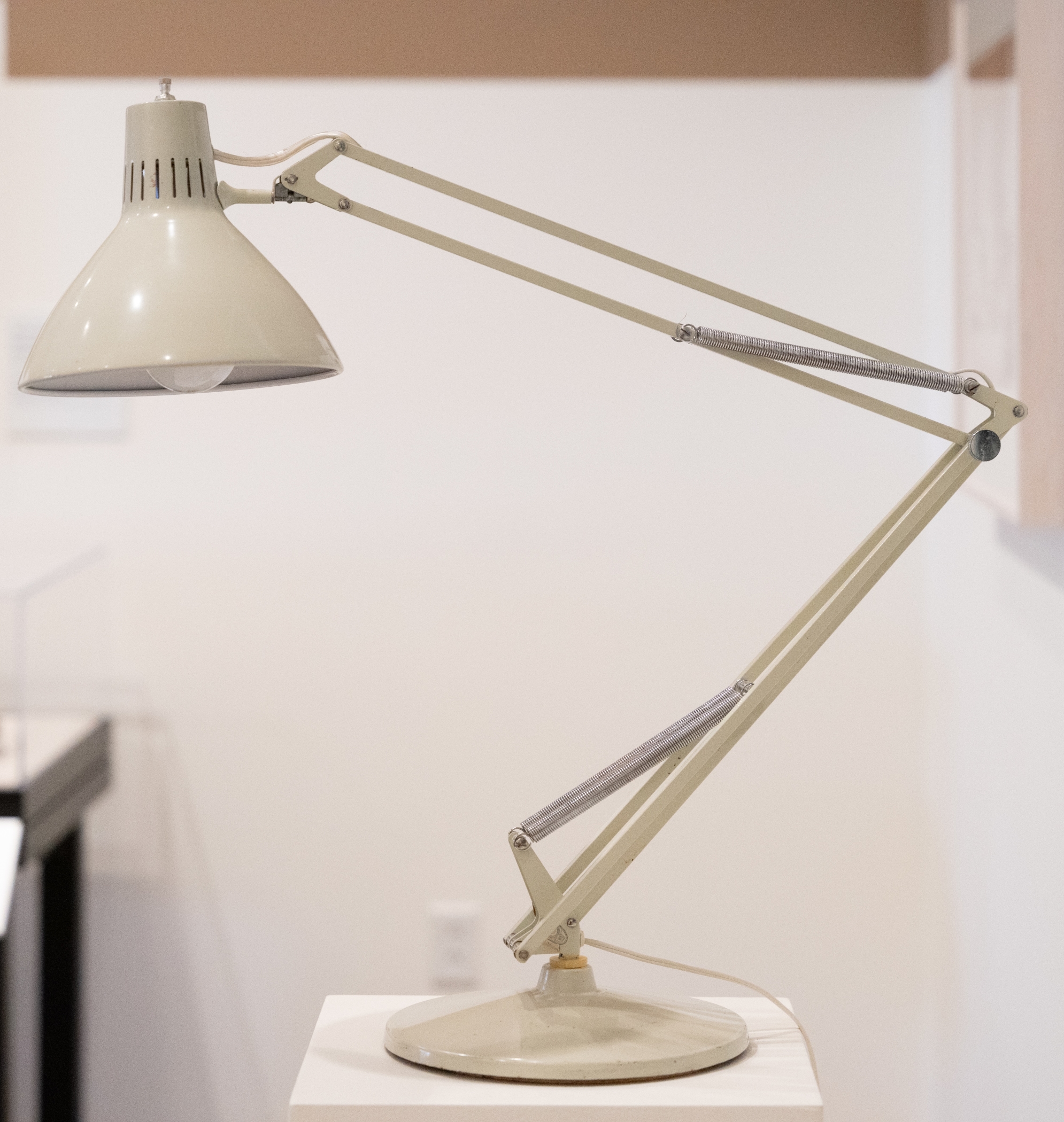 A simple cream-colored metal lampshade sits on an adjustable stem of the same color with silver springs and a rounded pedestal.