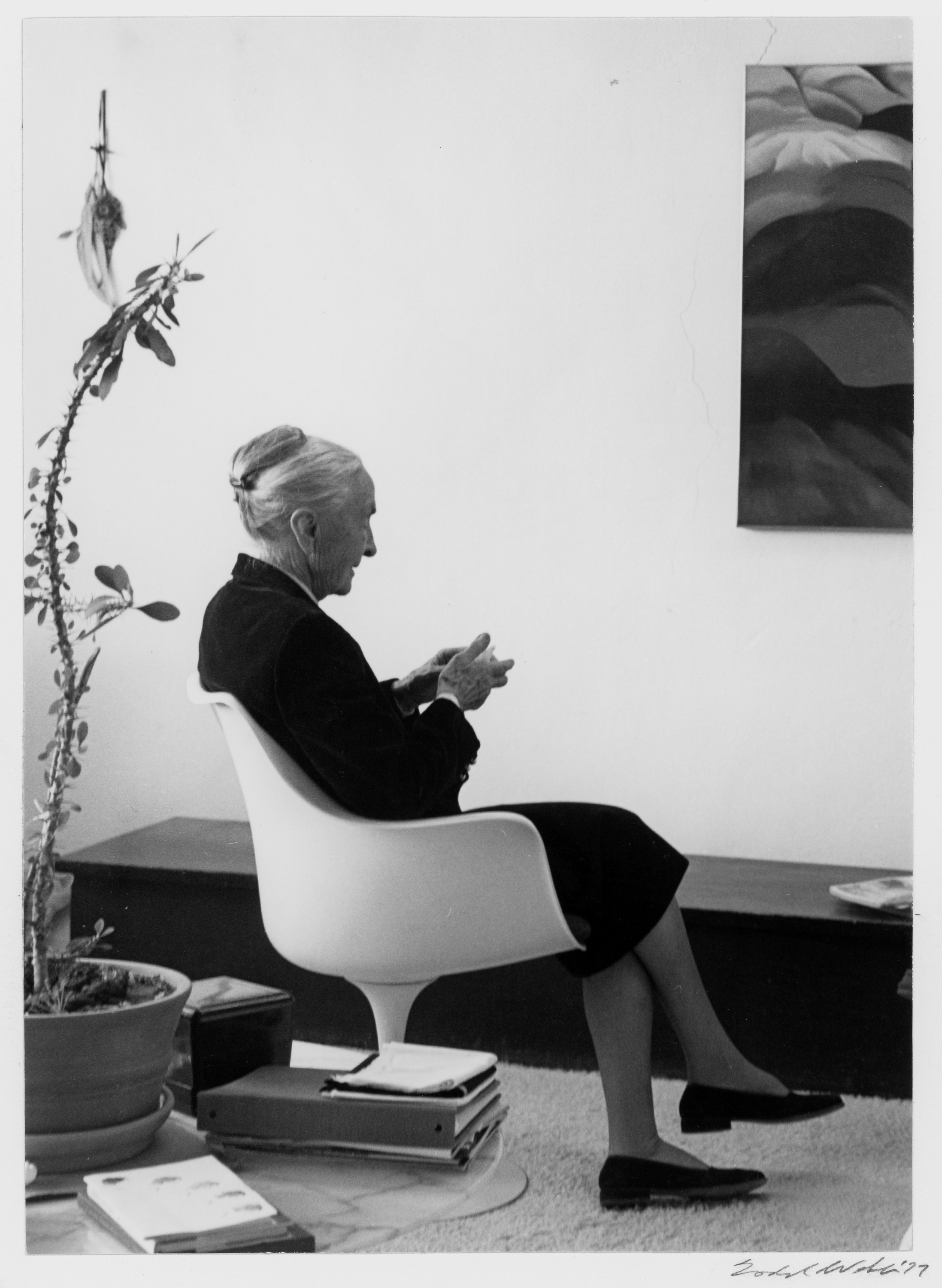 Georgia O'Keeffe with a gray bun and dark black dress is viewed from the side to feature a white molded chair that seems to hug her body. The woman holds her hands together and has her legs crossed. A tall skinny plant is behind her with a slight edge of a painting on the wall.