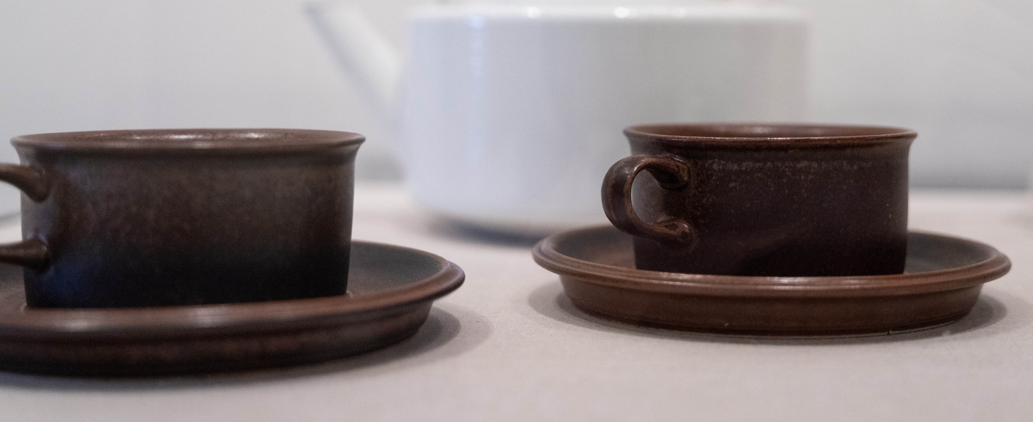 Two short dark brown mugs with subtle curves at the top sitting on matching saucers.