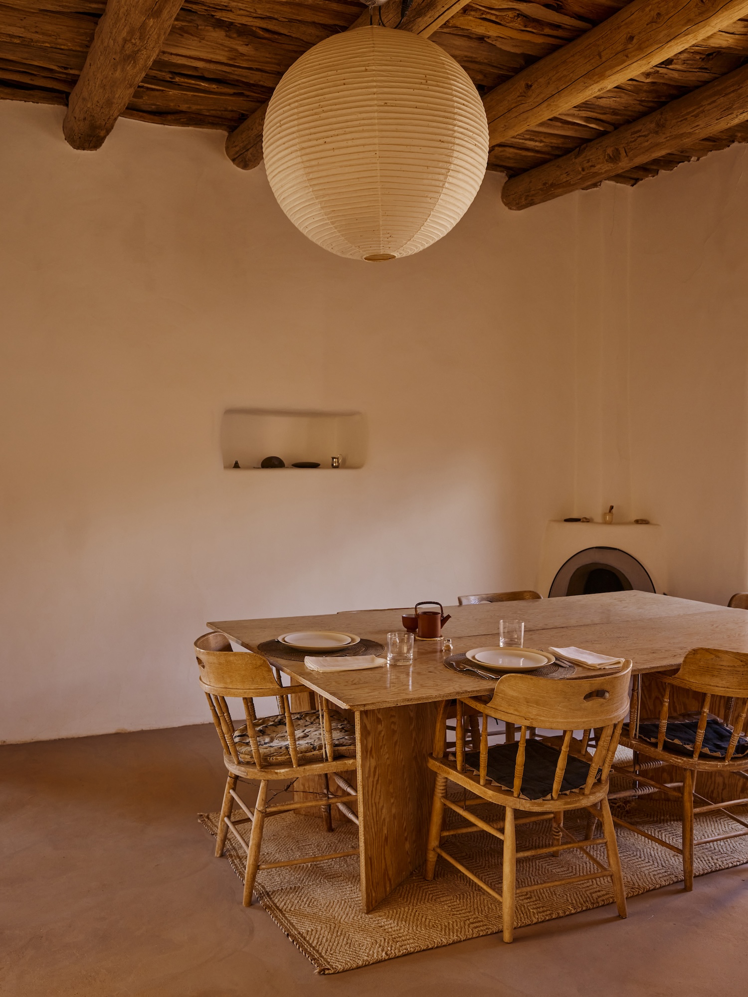 A dining room setting features a plywood table with rounded arm chairs made of wood tucked under the table. Above is a large globe-shaped light made of cream paper with horizontal stripes revealing the structure. The room has a built in shelf, or 'nicho' and a built in fireplace in the far corner.