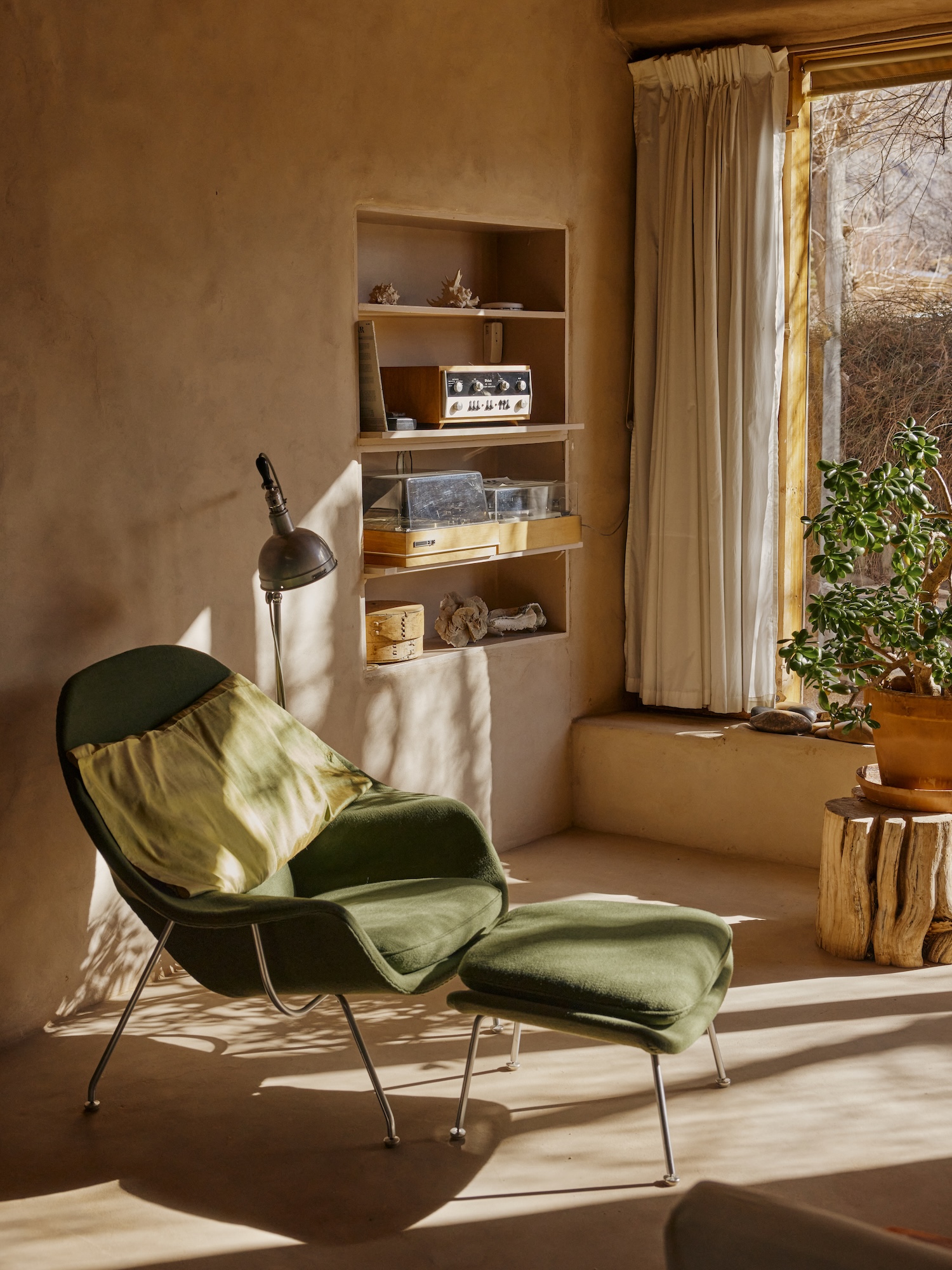 A living room setting featuring a green upholstered armchair with a matching ottoman with a yellow pillow against the back. A brown light sits next to the chair. Built-in shelves in the tan walls feature a stereo and other sparse items. A jade plant sits againsta a window in the background providing dappled light and shadow on the chair.