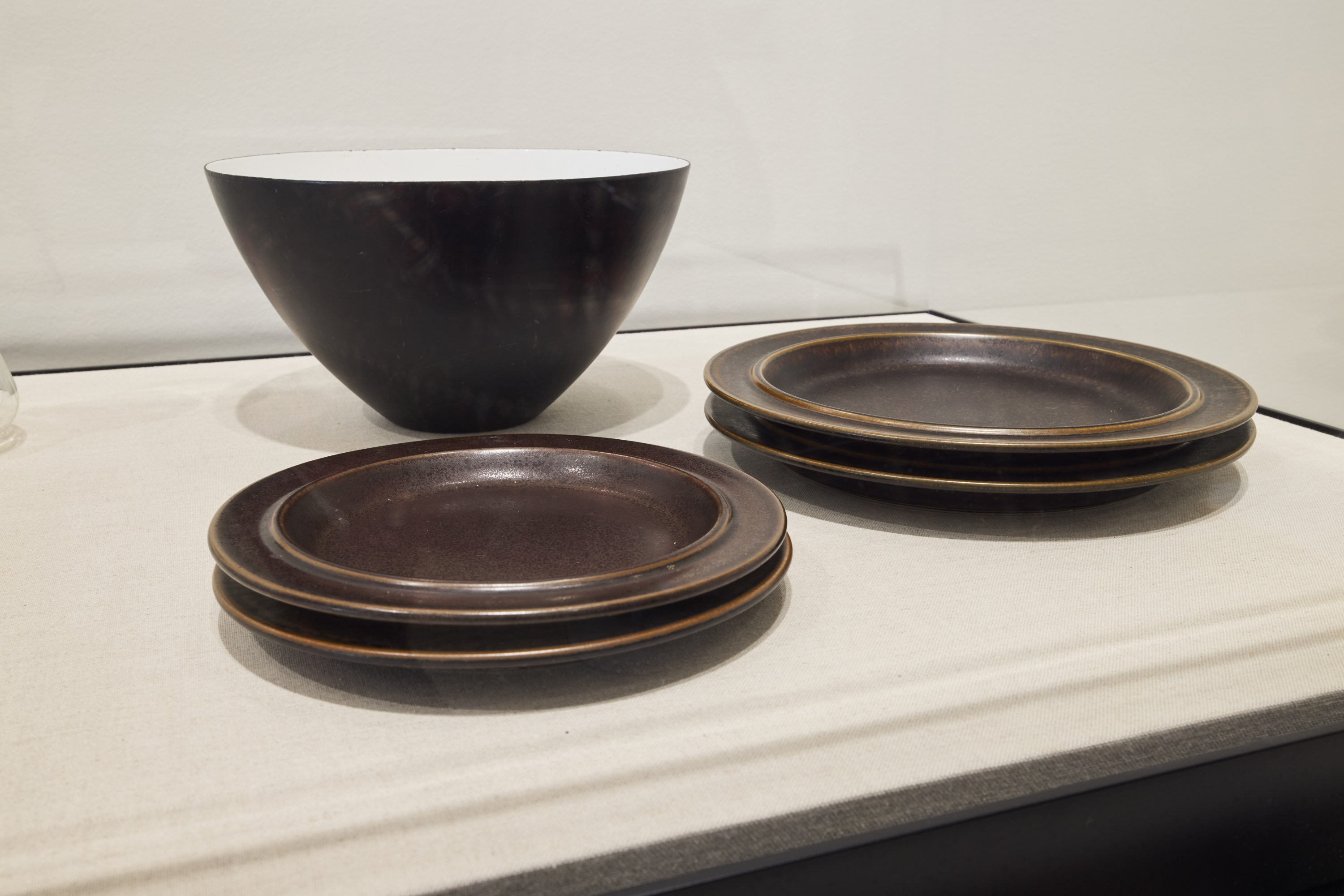 A dark brown tall salad bowl with a white interior. In the foreground are 2 stacks of 2 dark brown ceramic plates.