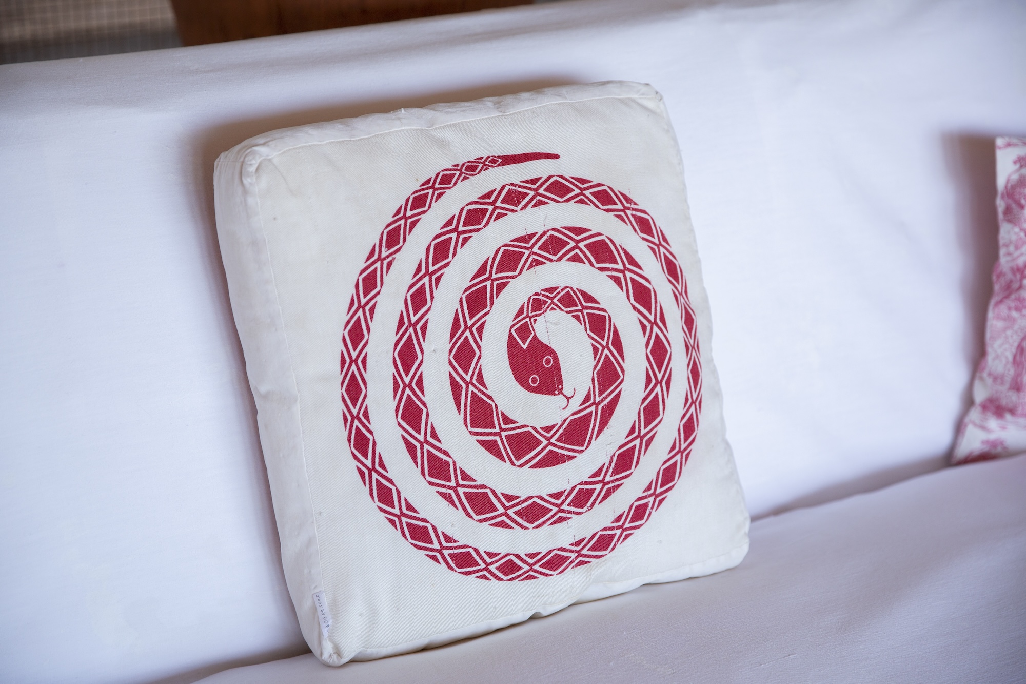 A white pillow with a graphic of a red snake in a spiral pattern with the snake's head at the center and tail on the outer edge. The pillow sits on a white background which seems to be a sofa.