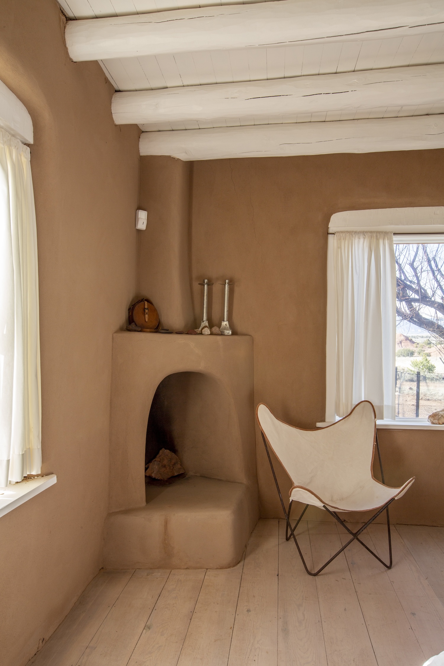 A chair made with a cream-colored canvas bordered in dark tan resembling the shape of a butterfly held together by a thin crossed pattern of black metal. The chair sits between a window and an built-in fireplace with a similarly dark tan color. The ceiling is painted white.